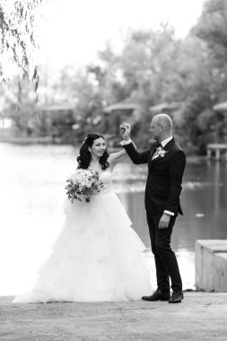 Beyaz elbiseli siyah saçlı bir gelin ve düğün günlerinde parkta papyonlu mavi elbiseli bir damat.