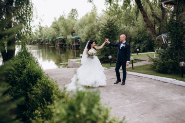 Beyaz elbiseli siyah saçlı bir gelin ve düğün günlerinde parkta papyonlu mavi elbiseli bir damat.