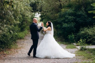 Beyaz elbiseli siyah saçlı bir gelin ve düğün günlerinde parkta papyonlu mavi elbiseli bir damat.