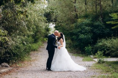 Beyaz elbiseli siyah saçlı bir gelin ve düğün günlerinde parkta papyonlu mavi elbiseli bir damat.