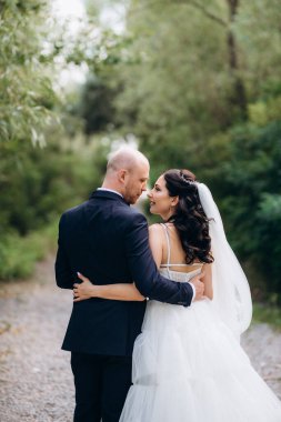 Beyaz elbiseli siyah saçlı bir gelin ve düğün günlerinde parkta papyonlu mavi elbiseli bir damat.