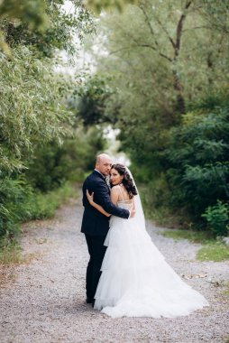 Beyaz elbiseli siyah saçlı bir gelin ve düğün günlerinde parkta papyonlu mavi elbiseli bir damat.