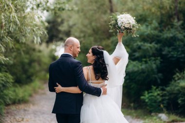 Beyaz elbiseli siyah saçlı bir gelin ve düğün günlerinde parkta papyonlu mavi elbiseli bir damat.