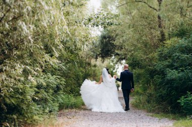 Beyaz elbiseli siyah saçlı bir gelin ve düğün günlerinde parkta papyonlu mavi elbiseli bir damat.