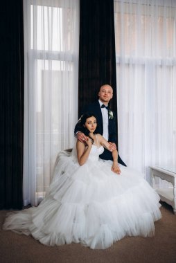 Beyaz elbiseli siyah saçlı bir gelin ve koyu renk bir otel odasında papyonlu mavi takım elbiseli bir damat.