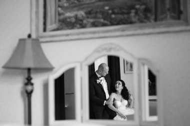 Beyaz elbiseli siyah saçlı bir gelin ve koyu renk bir otel odasında papyonlu mavi takım elbiseli bir damat.