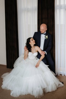 Beyaz elbiseli siyah saçlı bir gelin ve koyu renk bir otel odasında papyonlu mavi takım elbiseli bir damat.