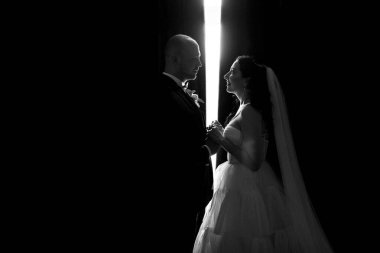 Beyaz elbiseli siyah saçlı bir gelin ve koyu renk bir otel odasında papyonlu mavi takım elbiseli bir damat.