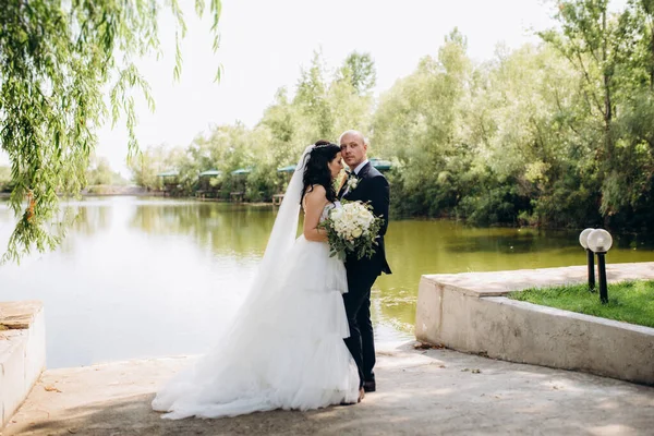 Beyaz elbiseli siyah saçlı bir gelin ve düğün günlerinde parkta papyonlu mavi elbiseli bir damat.