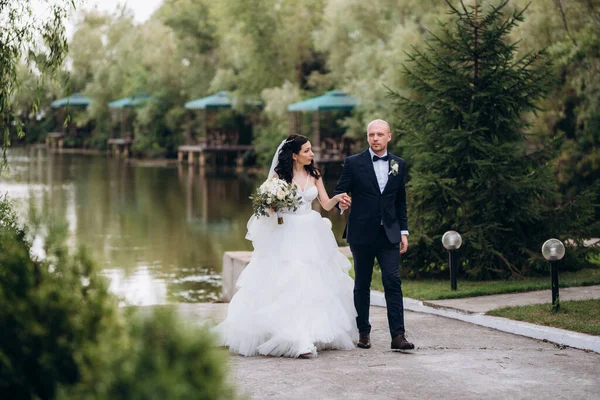 Beyaz elbiseli siyah saçlı bir gelin ve düğün günlerinde parkta papyonlu mavi elbiseli bir damat.