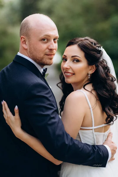 Beyaz elbiseli siyah saçlı bir gelin ve düğün günlerinde parkta papyonlu mavi elbiseli bir damat.