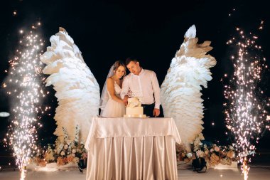 Düğün pastasını beyaz kanatların arka planına ve karanlık bir gece göğüne kesiyor.