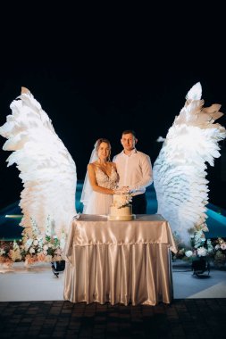 Düğün pastasını beyaz kanatların arka planına ve karanlık bir gece göğüne kesiyor.