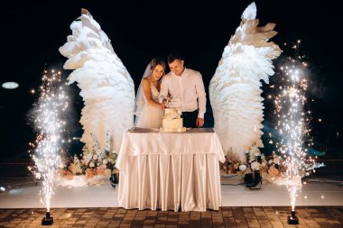 Düğün pastasını beyaz kanatların arka planına ve karanlık bir gece göğüne kesiyor.