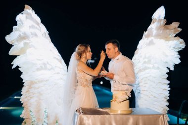 Düğün pastasını beyaz kanatların arka planına ve karanlık bir gece göğüne kesiyor.