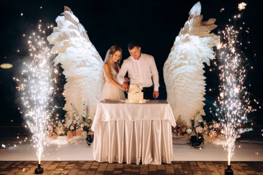 Düğün pastasını beyaz kanatların arka planına ve karanlık bir gece göğüne kesiyor.