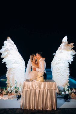 Düğün pastasını beyaz kanatların arka planına ve karanlık bir gece göğüne kesiyor.