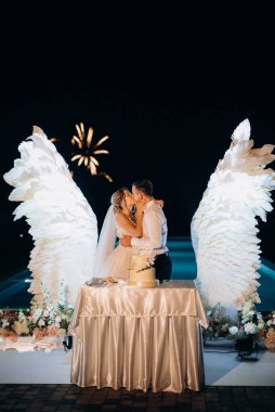 Düğün pastasını beyaz kanatların arka planına ve karanlık bir gece göğüne kesiyor.