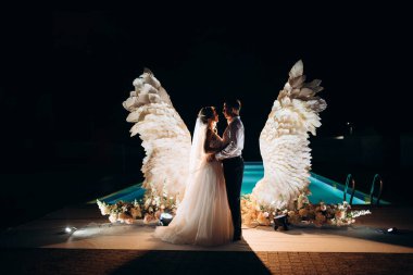 Düğün pastasını beyaz kanatların arka planına ve karanlık bir gece göğüne kesiyor.