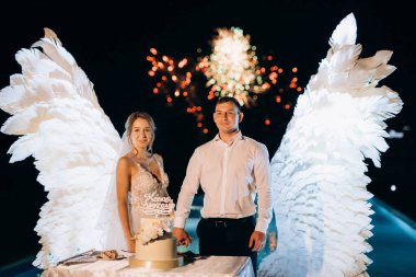 Düğün pastasını beyaz kanatların arka planına ve karanlık bir gece göğüne kesiyor.