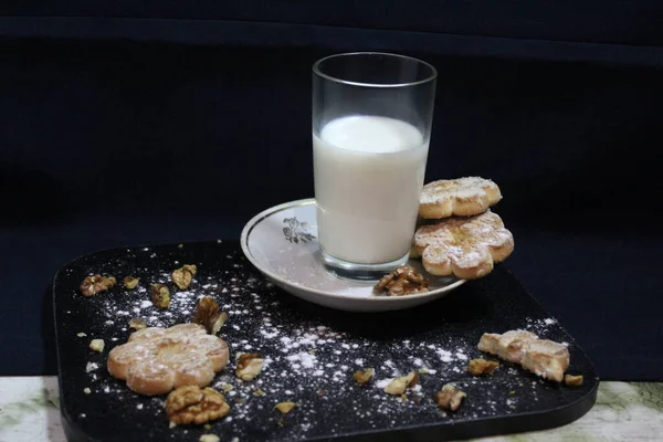 Karanlık bir tahtanın üzerinde bir tabak süt var. Arka planda kurabiye ve ceviz var. Durgun hayat toz şekerle serpiştirilmiş