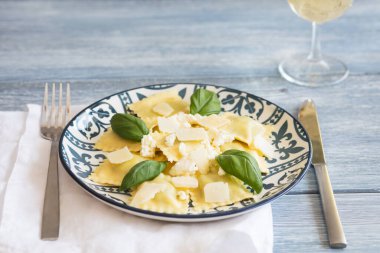 İtalyan desenli siyah bir arka planda parmesan serpiştirilmiş ricotta ve fesleğenli ravioli, bez peçetede çatal ve bir bardak beyaz şarap.