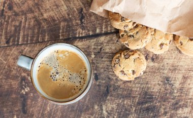 Çikolata parçalı kurabiyeler ve bir fincan kahve. Ahşap arka planda el işi torbası.