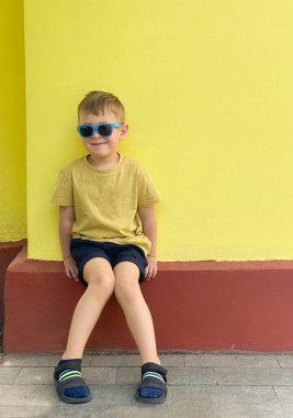 Parlak sarı bir arka planda, güneş gözlüklü, gülümseyen küçük çocuk.