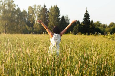 Arkadan siyah saçlı genç bir kadın açık kollarla tarlada duruyor.