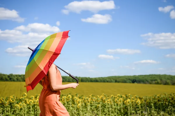 Joyful beautiful girl holding multicolored umbrella in sunflower field and blue cloud sky background — Stock Photo, Image