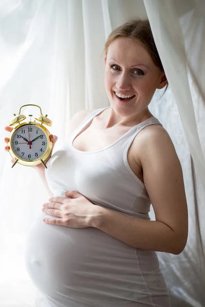 Holding alarm clock pregnant beautiful young lady on light window copy space background — Stock Photo, Image