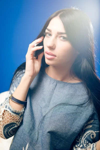 Фотографія красивої молодої жінки з розкішним волоссям, що розмовляє на мобільному телефоні і чуттєво дивиться на камеру на синьому копіювальному просторі — стокове фото