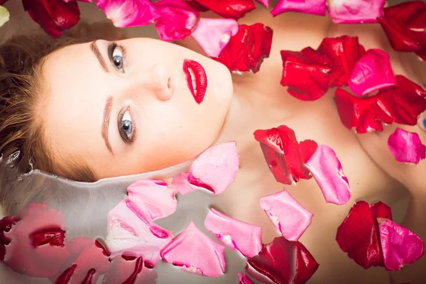 Luxury spa: elegant blue eyes lady pinup girl with silk skin having fun happy relaxing laying in water bath & looking at camera on colorful rose flower petals copy space background close up portrait — 스톡 사진