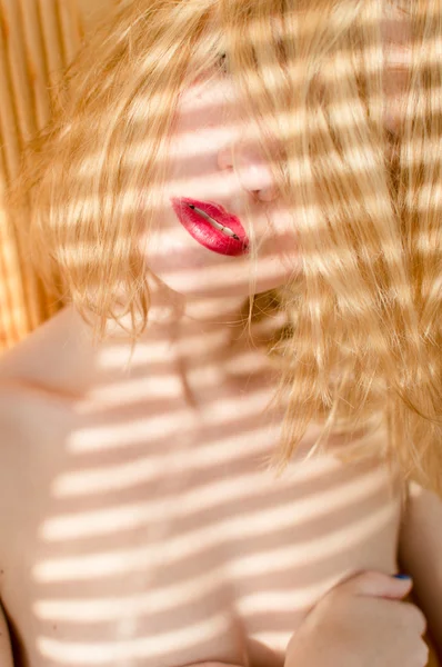 Крупним планом на червоній помаді блондинка молода красива леді піпп дівчина весело позує, розслабляючись з тіні від сонячних променів через віконні жалюзі, що стоять біля стіни і дивлячись на портрет камери — стокове фото