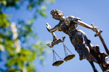 back of sculpture themis, femida or justice goddess on bright blue sky and green leaves outdoors background