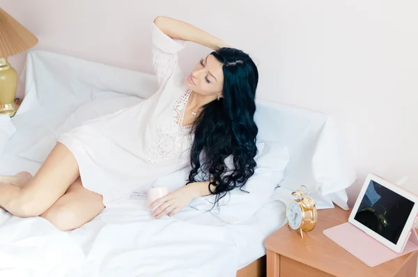 Портрет чудової сексуальної красивої дівчини у відмінній формі, яка весело розслабляється, лежачи в ліжку і позує з ноутбуком та будильником на світлому фоні копіювання простору — стокове фото