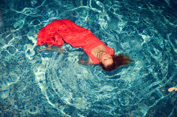 Seductive female floating on swimming pool in gorgeous red dress. — Stockfoto