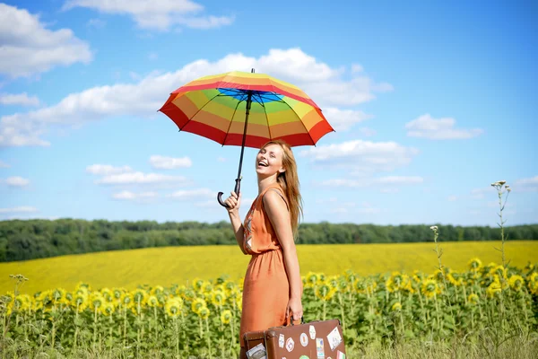 Портрет красивой молодой блондинки с чемоданом для путешествий и радужным зонтиком на подсолнечном поле на открытом воздухе — стоковое фото