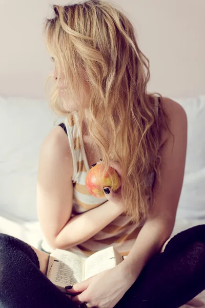 Picture of blonde sexi female having fun happy holding book and big red apple sitting on white bed on light background and hiding face