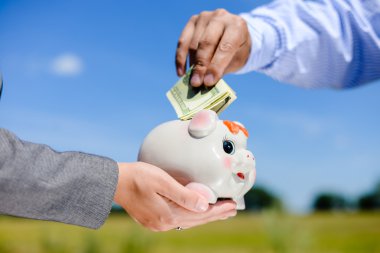 Picture of hands holding a piggy bank with a dollar bills stuck in the top slot. Side view in horizontal format over a blue sky sunny outdoors background copy space.