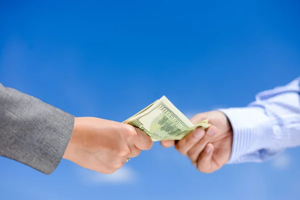 Closeup of of hands giving and receiving dollar bills. Side view in horizontal format over a blue sky sunny outdoors, background copy space