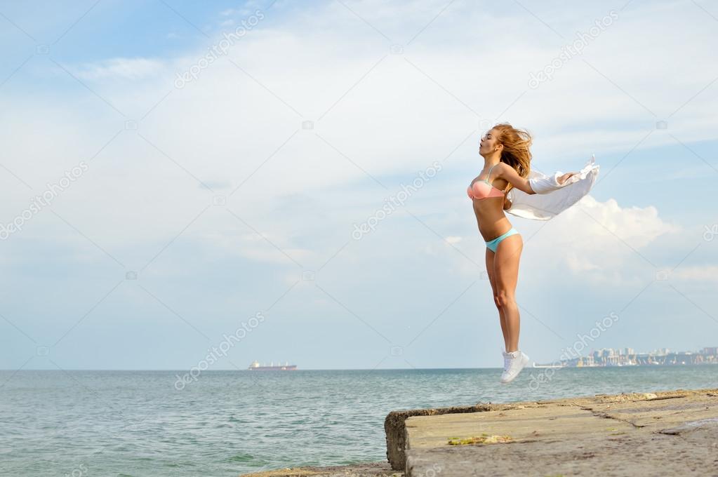 Young lady with arms extended sidewards jumping up on rocky beach 