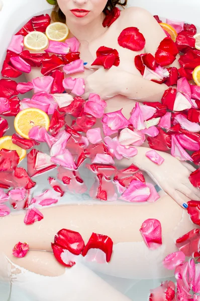 Красива сексуальна елегантна молода леді з червоною помадою та шовковою шкірою весело лежить у водяній бані, розслабляючись на барвистому фоні пелюсток троянд — стокове фото