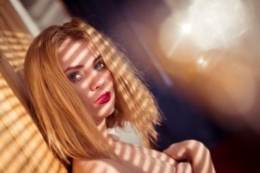 close up on elegant pretty lady with shadow from window blinds on bare shoulders having fun looking at camera surrounded by sun light flare rays on copy space background