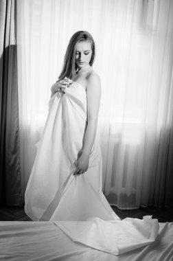 Black and white portrait of young beautiful lady hiding under white sheet standing on light window copy space background 