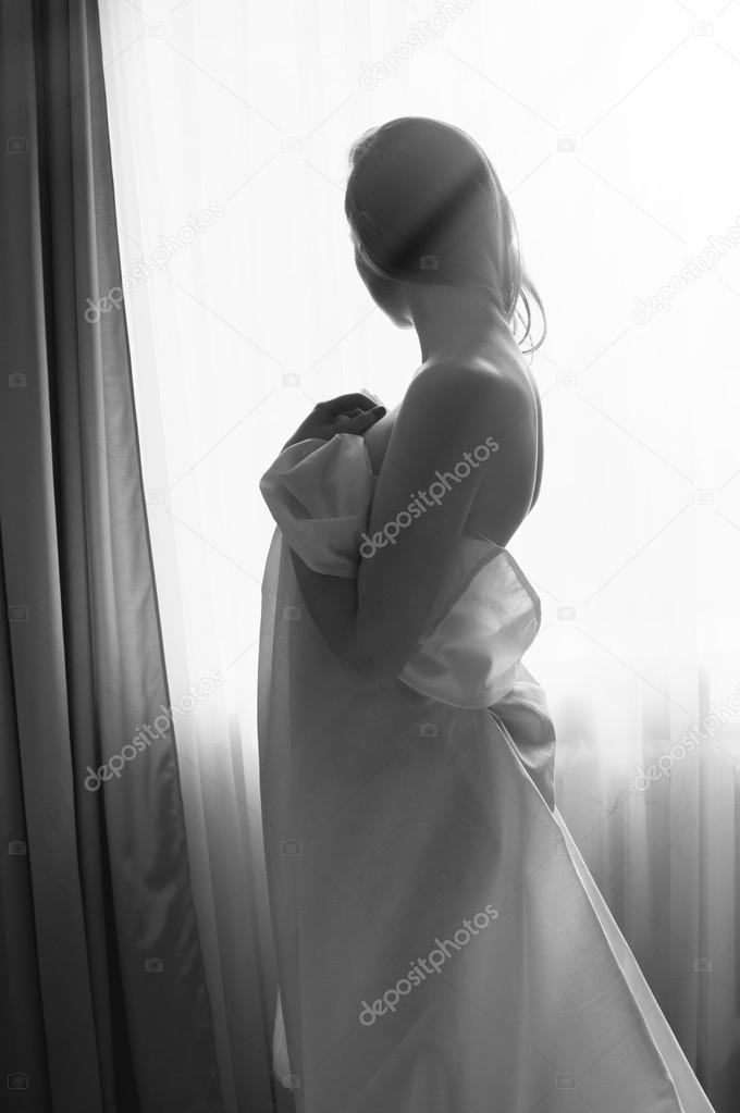 Blond young beautiful woman having fun hiding under white sheet standing on light window copy space background. Black and white portrait 