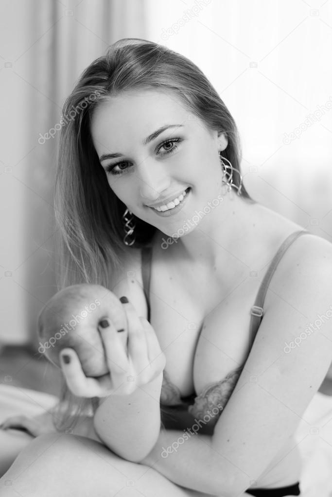 Sexy blonde girl having fun happy smiling holding big apple sitting on bed in lingerie. Black and white portrait 