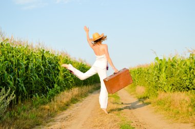 Jumping girl wearing hat with suitcase on road in field over blue sky outdoors background