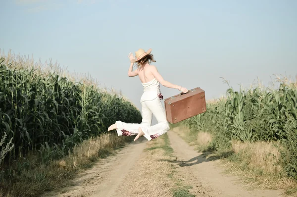 Jumping girl wearing straw hat with suitcase in corn field — Stockfoto