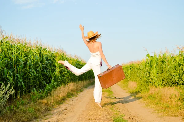 Jumping girl wearing hat with suitcase on road in field over blue sky outdoors background — Stock Photo, Image